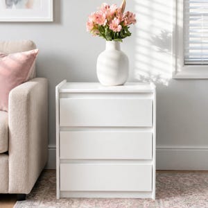 May include: A white three-drawer nightstand with a smooth finish and clean lines. A white ceramic vase sits on top of the nightstand, filled with a bouquet of pink alstroemeria flowers. The piece features a simple, rectangular design.
