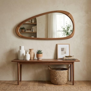 May include: A large, organically shaped wooden mirror hangs above a dark wood console table. The table displays white and beige vases, a potted succulent, and a framed print. A woven basket sits below. The room has a neutral color palette.