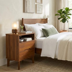 May include: A wooden nightstand with an open shelf and two drawers, topped with a cylindrical lamp. The nightstand sits beside a bed featuring a wooden headboard, white bedding, a green throw pillow, and a potted fiddle leaf fig plant.