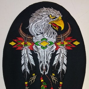 May include: An embroidered patch with a white eagle head, a skull with horns, and feathers in red, green, yellow, and white colors on a black background.