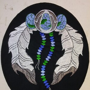 May include: A black embroidered patch with a white and gray feather design, a blue and green beaded string, and a silver and blue circular design in the center.