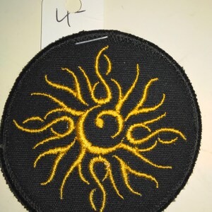 May include: A black circular patch with a gold embroidered sun design. The sun has a stylized eye in the center.