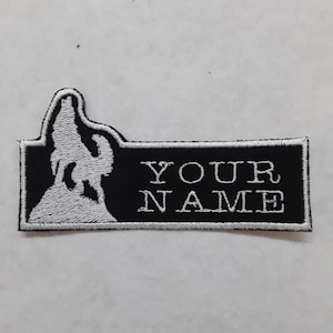 May include: Black and white embroidered patch featuring a howling wolf silhouette on the left side. The patch has the text "YOUR NAME" in white, centered on a black rectangular background. The patch is rectangular in shape.
