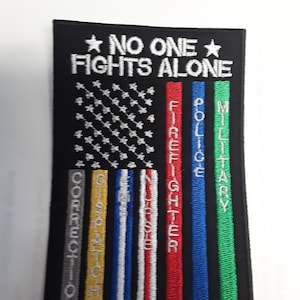 May include: Black embroidered patch with a white American flag design. The flag has a thin red, white, and blue stripe on the right side. The text "NO ONE FIGHTS ALONE" is above the flag. The text "FIREFIGHTER", "POLICE", "MILITARY", "DISPATCH", and "CORRECTIONS" are written vertically on the stripes.