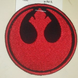 May include: A red embroidered patch with a black Rebel Alliance symbol. The symbol is a stylized bird's wing with a central point.