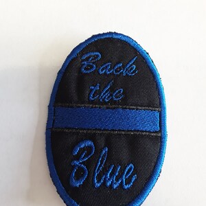 May include: A black oval patch with blue embroidered edging. The patch has a blue stripe across the center and the words "Back the Blue" embroidered in blue.