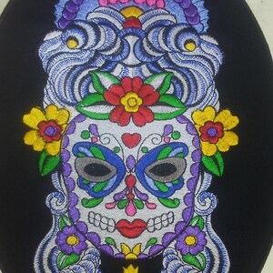 May include: Embroidered patch featuring a colorful sugar skull with a butterfly design, flowers, and a cross. The skull is white with black outlines and features a red mouth, blue eyes, and a pink nose. The background is black.
