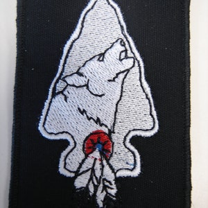 May include: Black rectangular patch with a white embroidered arrowhead design. The arrowhead features a howling wolf and a feather with red and blue accents. The patch is likely for clothing or accessories.