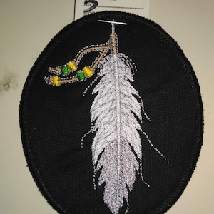 May include: A black oval patch with a white feather and brown and green beads. The patch is embroidered with a white feather and brown and green beads.
