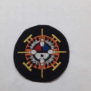 May include: Black and orange embroidered patch with a circle design featuring a white, red, and blue circle in the center, surrounded by feathers and four gold bars.