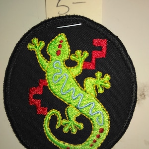 May include: A green lizard with red and blue details is embroidered on a black circular patch. The patch has a black border.