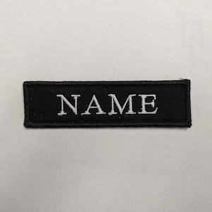 May include: Black fabric patch with white embroidered text that reads "NAME".