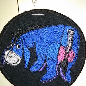 May include: A blue embroidered patch of a cartoon donkey with a pink bow on its tail. The donkey is on a black background.