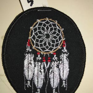 May include: Black oval fabric patch with an embroidered dreamcatcher design. The dreamcatcher has a white and gold web with red beads and five white feathers hanging from the bottom.