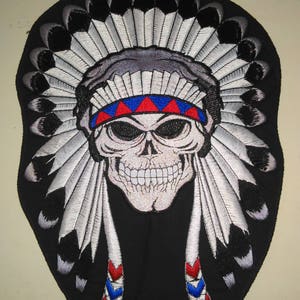 May include: Embroidered iron-on patch featuring a white skull wearing a black and white headdress with red and blue accents.