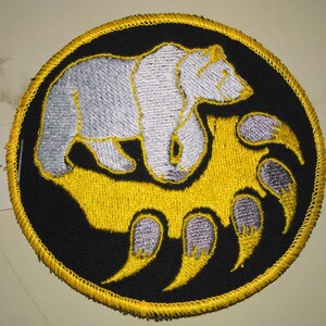 May include: A round black patch with a yellow border. A white polar bear is walking on top of a yellow bear paw print with gray claws.