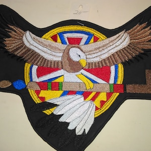 May include: Embroidered patch featuring a brown eagle with outstretched wings, holding a peace pipe in its talons. The eagle is centered in a circular design with red, blue, yellow, and green colors. The patch is on a black background.