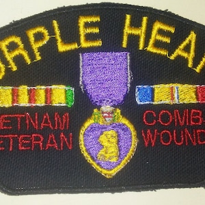 May include: A black fabric patch with the words "Purple Heart" in yellow embroidery. The patch also features a purple heart medal, a Vietnam Veteran ribbon, and a Combat Wounded ribbon. The words "Vietnam Veteran" and "Combat Wounded" are in red embroidery.