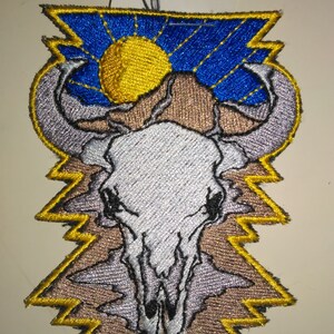 May include: An embroidered patch featuring a white animal skull with a yellow sun and blue sky in the background. The skull is surrounded by a brown and yellow zig-zag pattern.