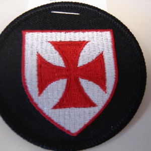 May include: A round black patch with a white shield and a red cross in the center. The patch is embroidered.