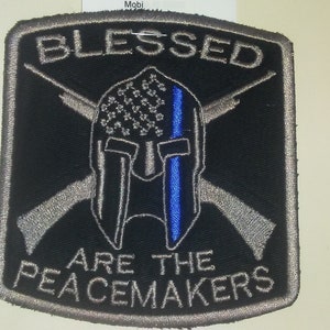 May include: A black patch with a silver embroidered border. The patch features a silver Spartan helmet with a blue and white American flag design. Two silver rifles are crossed behind the helmet. The text "BLESSED ARE THE  PEACEMAKERS" is embroidered in silver.