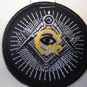 May include: A black embroidered patch with a white and silver all-seeing eye inside a triangle, surrounded by a ring of white rays. The eye is looking directly at the viewer. The triangle is surrounded by a square with a compass and a level on each side. The patch is circular and has a black border.