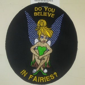 May include: A black and yellow embroidered patch with a cartoon fairy sitting on a mushroom. The fairy has green wings, a green dress, and blonde hair. The text "Do you believe in fairies?" is written in yellow around the fairy.