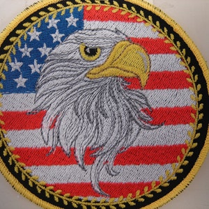 May include: Embroidered patch featuring a bald eagle's head against a backdrop of the American flag. The eagle has detailed gray and white feathers, a yellow beak, and a focused gaze. The patch is circular, with a black and gold border.