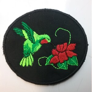 May include: A green hummingbird with a red throat is perched on a red flower with green leaves. The image is embroidered on a black circular patch.