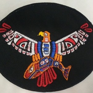May include: A black oval patch with an embroidered design of a stylized eagle holding a fish in its talons. The eagle is red, white, blue, and orange. The fish is red, orange, blue, and white.