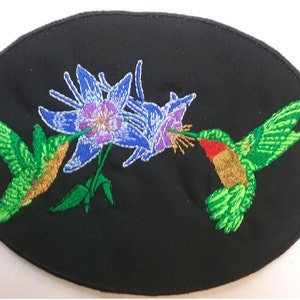 May include: Embroidered patch featuring two green hummingbirds with gold accents, one feeding from a purple and blue flower with green leaves on a black background.