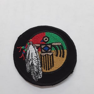 May include: A round black patch with a colorful embroidered design. The design features a feather, a circle with red, green, and blue sections, and a gold-colored symbol with vertical lines.