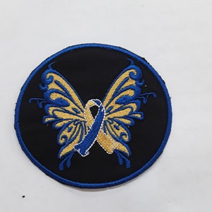 May include: A round black patch with a blue border features a gold butterfly with blue and gold ribbon wings.
