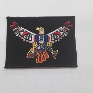 May include: A black embroidered patch featuring a stylized eagle with outstretched wings holding a fish in its talons. The eagle is depicted in red, white, blue, yellow, and orange colors.