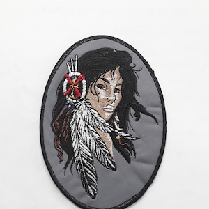 May include: Embroidered patch featuring a Native American woman with long black hair, wearing a beaded headdress with feathers. The patch is oval-shaped and has a gray background.