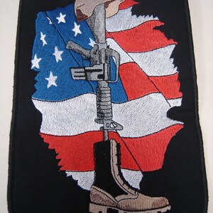May include: A black embroidered patch with a tattered American flag, a rifle, a military boot, and a helmet. The rifle is standing upright with the boot at the bottom and the helmet at the top. The flag is behind the rifle and boot.
