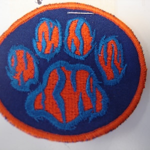 May include: A round, orange and blue embroidered patch with a paw print design. The paw print is outlined in blue and filled with orange and blue stripes.