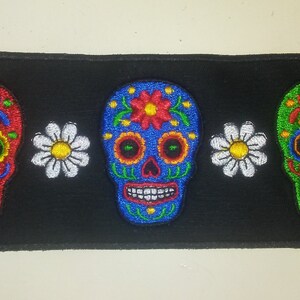 May include: Three embroidered sugar skull patches on a black background. The skulls are red, blue, and green with white flowers on either side.