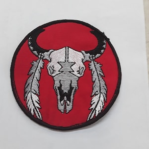 May include: A red embroidered patch with a white and black buffalo skull with feathers on either side. The patch is circular and has a black border.