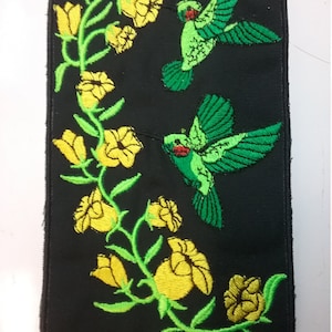 May include: Embroidered patch featuring two green hummingbirds with red beaks flying near yellow flowers on a black background.