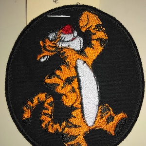 May include: A black and orange embroidered patch featuring a cartoon tiger with a white belly and a red nose. The tiger is standing on its hind legs with its front paws raised in the air. The patch is circular and has a black border.