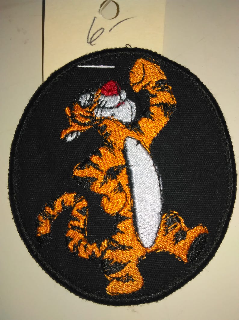 May include: A black and orange embroidered patch featuring a cartoon tiger with a white belly and a red nose. The tiger is standing on its hind legs with its front paws raised in the air. The patch is circular and has a black border.