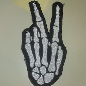 May include: A black and white embroidered patch of a skeleton hand making the peace sign.