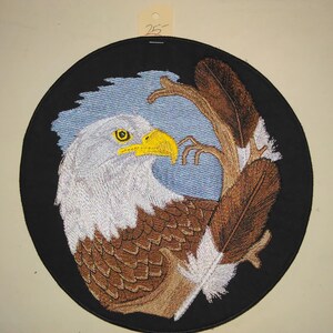 May include: Embroidered patch featuring a detailed bald eagle design. The eagle has a white head, brown body, and yellow beak, set against a blue sky and brown branch, all within a black circular border. A tag with the number 25 is attached.
