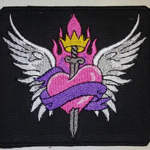 May include: Embroidered patch with a pink heart pierced by a sword with a gold crown on top. The heart is wrapped in a purple ribbon and surrounded by white wings.