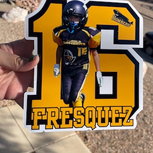 Puede incluir: Una pegatina troquelada con un jugador de fútbol americano con uniforme azul marino y dorado, con el número 16 y el nombre FRESQUEZ en letras amarillas. La pegatina también incluye el nombre del equipo "Bombers".