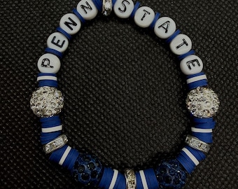 Può includere: Un braccialetto circolare di perline con lettere bianche che compongono la scritta PENN STATE, intervallate da perline piatte a disco blu e bianche, due sfere argentate incastonate di strass, due anelli distanziatori in cristallo e due sfere blu scuro testurizzate.