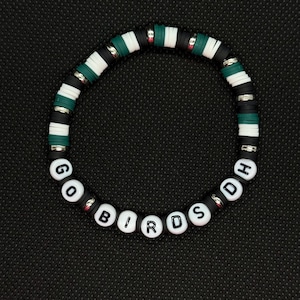 May include: A circular bracelet with alternating black, white, and green disc-shaped beads. The bracelet spells out "GO BIRDS" in white letter beads. The background is a textured black surface.
