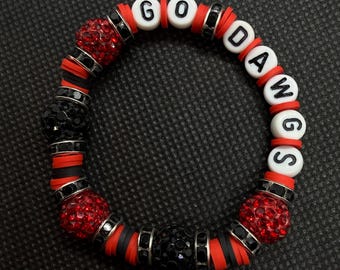 Può includere: Un braccialetto circolare di perline con lettere bianche che compongono la scritta GO DAWGS, intervallate da perline piatte a disco rosse e nere, perline sferiche scintillanti con strass rossi e neri e anelli distanziatori in metallo argentato.