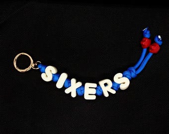 Può includere: Un portachiavi in paracord blu intrecciato con perline a lettera bianche che compongono la parola SIXERS. Il portachiavi include un anello in metallo dorato a un'estremità e due perline rosse a forma di stella sulle estremità del cordino.